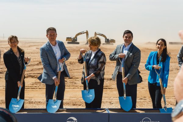 A row of people wearing suits hold up shovels at a construction site.