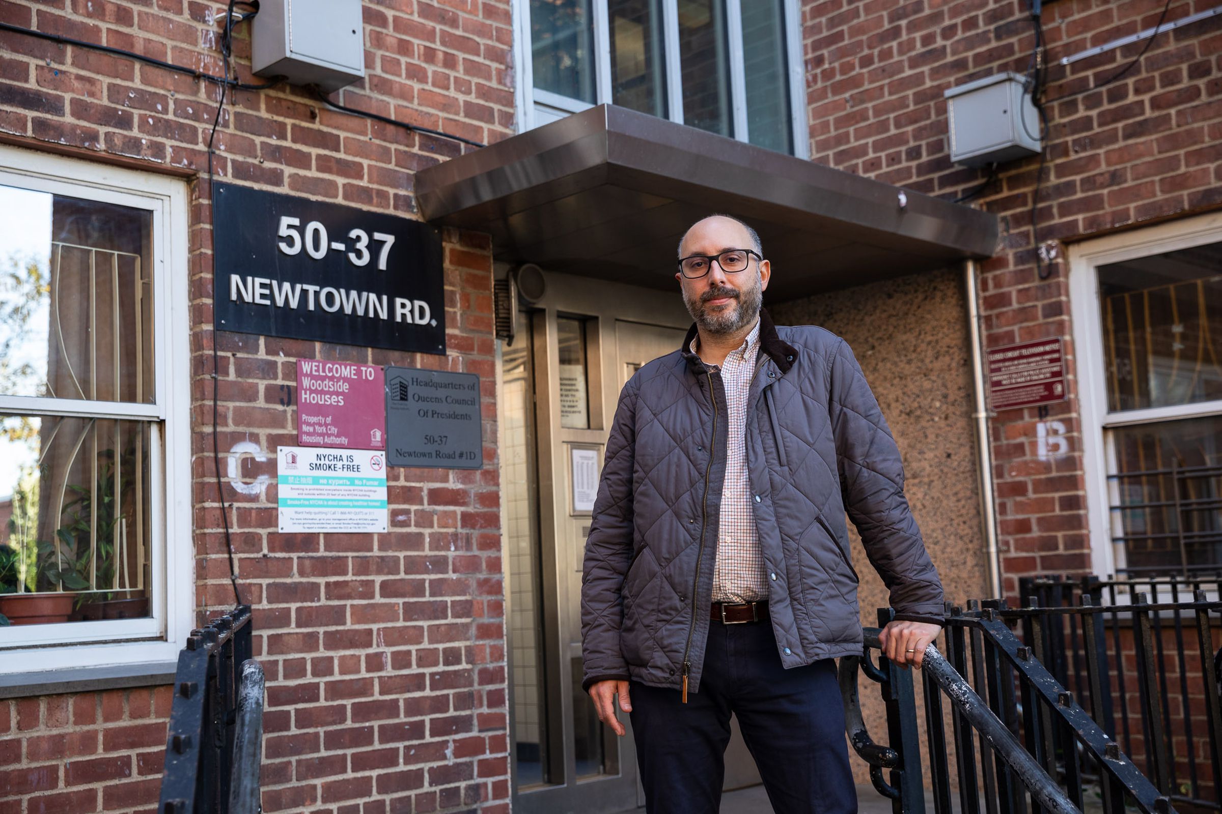 A man wearing a jacket and glasses stands in front of the door to an apartment building.